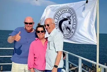 2b7517289cc5fe7734694eca33226758 Three people stand on a boat deck, with a "Fieldworkers Club" flag and the ocean in the background. One person gives a thumbs-up.