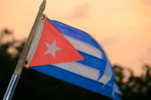 41b5b0790c56d90d4bd78ccb8f171d59 The Cuban flag waving against a peach-colored sky, featuring horizontal blue and white stripes, a red triangle, and a white star.