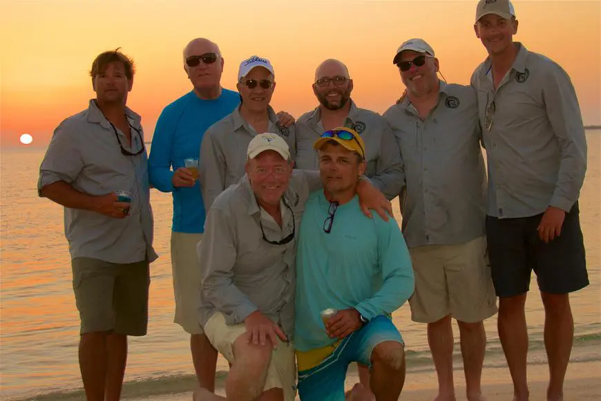 4bb9632ac1733cf6a440236d6bb74383 Eight men posing on a beach at sunset, wearing casual clothes and sunglasses. They are smiling and standing close together with the ocean and setting sun in the background.