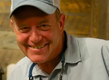 b2f4a95c2b050cba9edf1eab79f16bc7 Smiling man wearing a light blue cap and shirt, with sunglasses hanging from his neck. Wood-textured background.