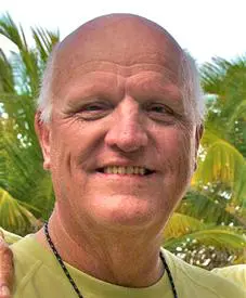c2c1ce87c062a1ed2e5f97f2b81f4a57 A smiling man with a bald head and wearing a green shirt stands in front of palm trees.